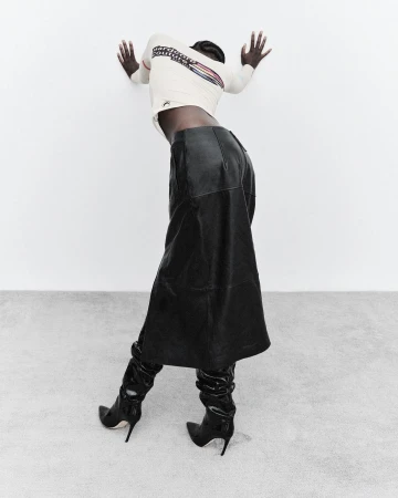 Fashion shot of a woman with dark hair in a white long-sleeve shirt, black leather skirt, and high-heeled boots against a white wall and floor.