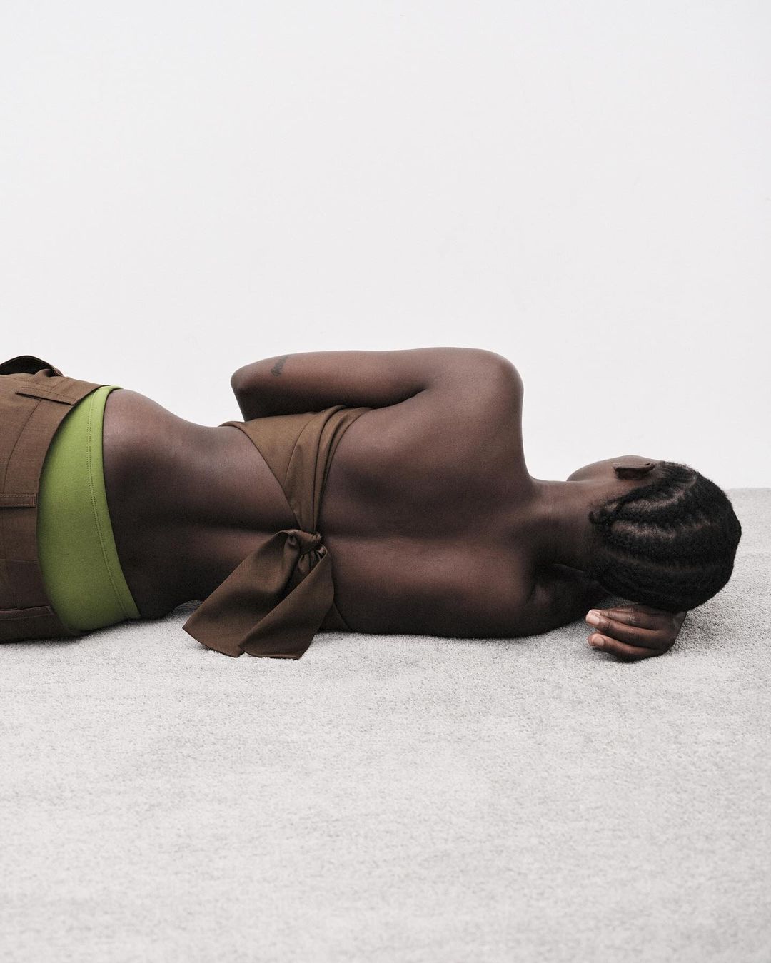 An adult woman lies on a grey surface wearing a brown top and green bottoms in a brightly lit, neutral-toned indoor setting.
