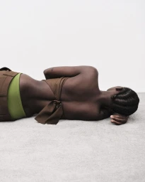 Black woman lying prone on grey floor wearing brown backless top and green waistband