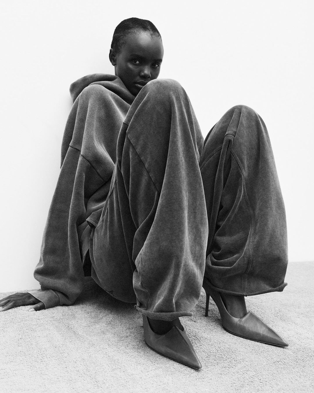 Woman in oversized hoodie and pants sits on carpet. She wears heels. Black and white fashion photograph on light grey background.