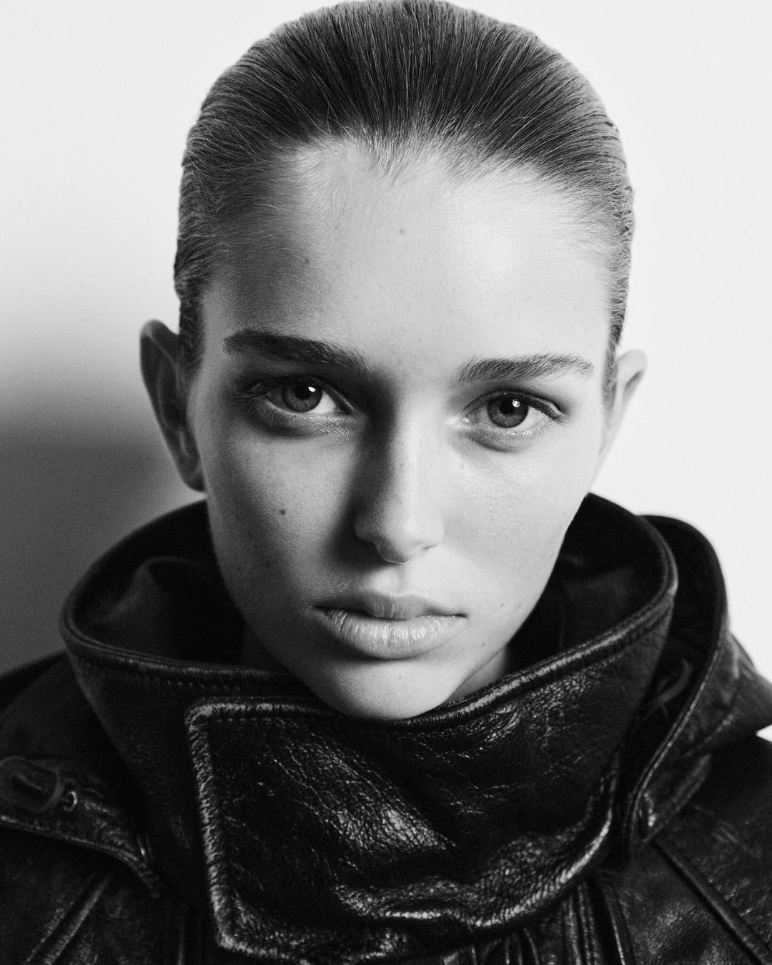 Black-and-white portrait of a woman with a slicked-back hairstyle wearing a black leather jacket, set against a bright background.