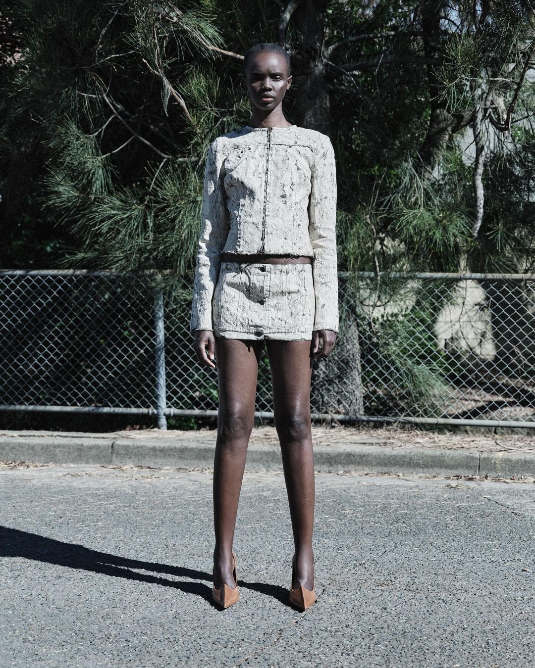 Model standing outdoors wearing a textured white patterned jacket and matching mini skirt against a wire fence and pine trees.