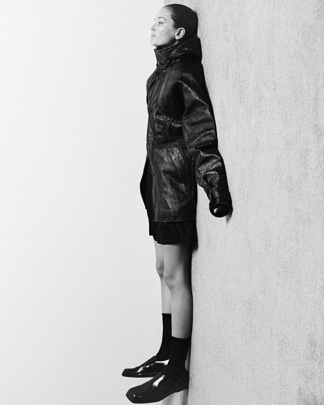 Woman in black coat leaning against wall, showcasing fashion with shiny shoes and dark socks in a studio with stark lighting.
