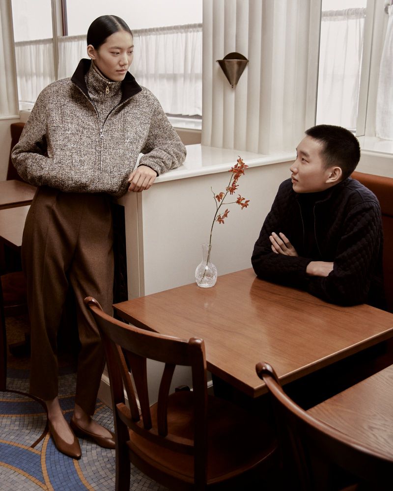 Two people in a cafe or restaurant setting. A woman in a wool sweater stands next to a table while a man in a sweater sits at the table.