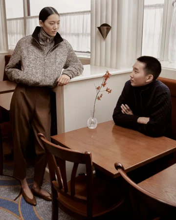 Two people in a cafe or restaurant setting. A woman in a wool sweater stands next to a table while a man in a sweater sits at the table.