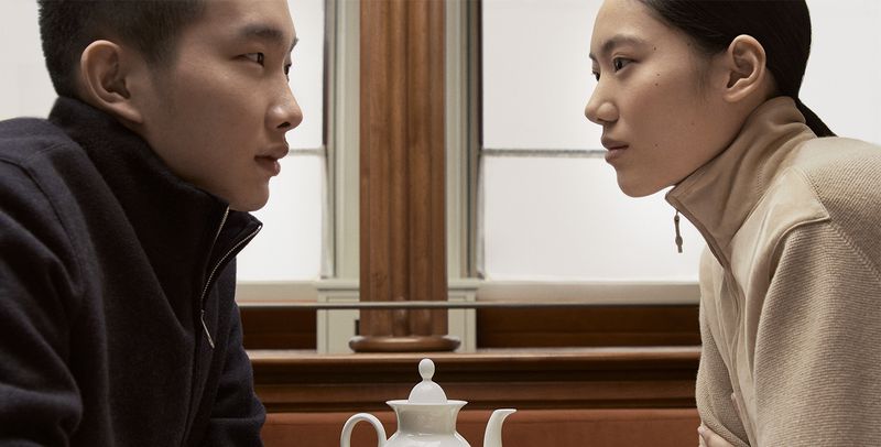 Portrait of an Asian man and woman facing each other indoors near a white teapot, with a neutral-color palette and studio lighting.