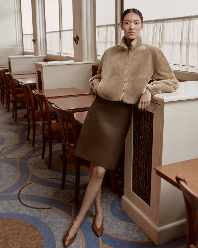 Adult woman in a tan jacket and brown skirt posing in a restaurant with tables and patterned floor.