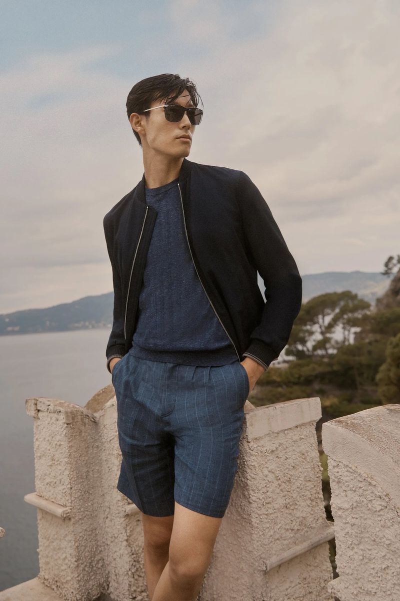 A stylish adult male poses outdoors wearing a blue shirt, blue shorts, and a black jacket with sunglasses on a sunny day.