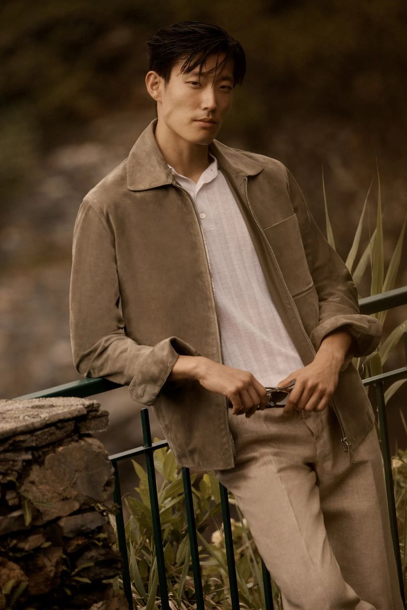 Man in neutral tones poses in a tan jacket and pants leaning against a stone railing in natural outdoor lighting, shot at eye level.