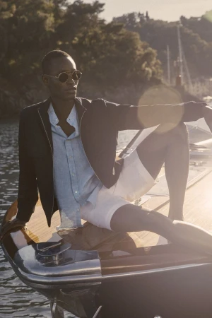 Model wearing dark sunglasses, dark jacket, light shirt and white shorts seated on a boat deck at sunset.
