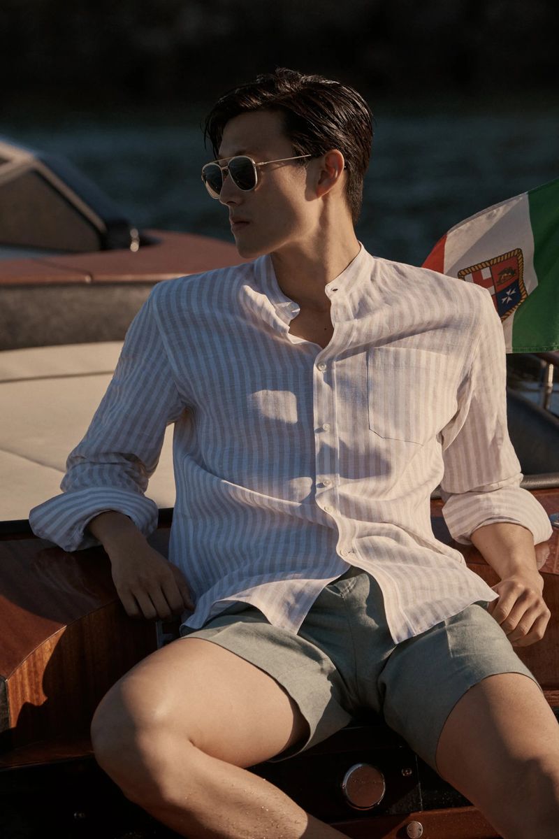 Adult male poses on a boat wearing sunglasses, a vertically striped shirt, and olive-green shorts with an Italian flag in background.