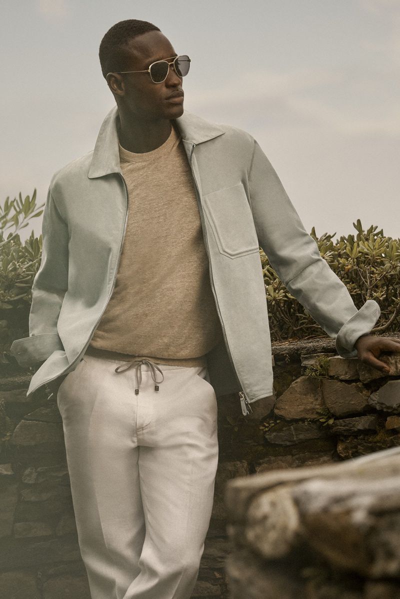 Adult male wearing aviator sunglasses, light jacket, and white pants poses against a stone wall outside on a sunny day.