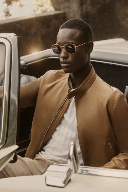 Portrait of an African American man in sunglasses and a tan jacket, seated in a vintage convertible car on an outdoor location.