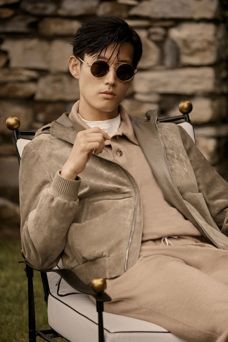 A stylish Asian man in round sunglasses and a beige leisure suit sits outside on a black and white chair with a stone wall backdrop.