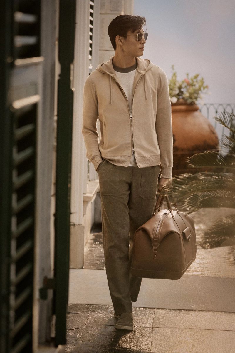 An Asian man walks outdoors wearing a hoodie, pants, and sunglasses, carrying a brown leather bag. Balustrades and greenery visible.