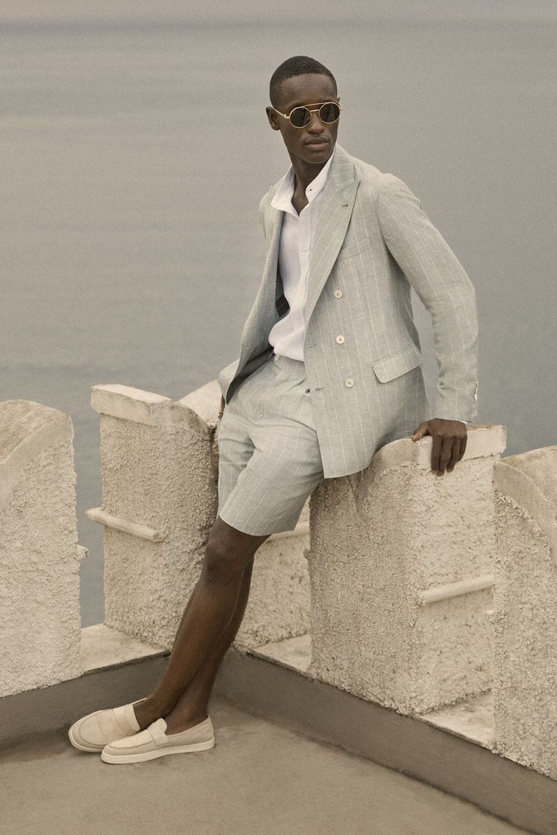 An African American male model poses in a light grey suit with shorts and round sunglasses on a stone ledge with the sea in the background.
