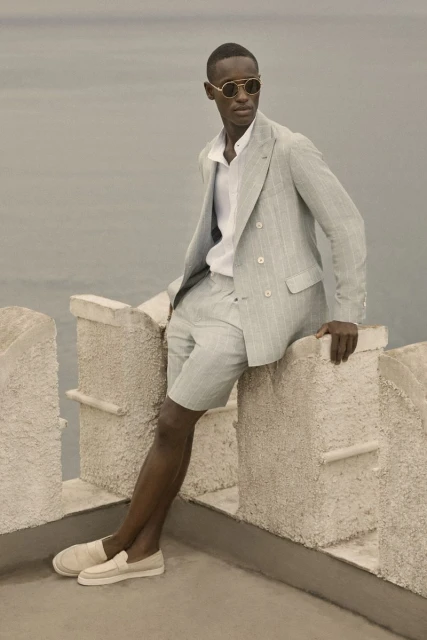 An African American male model poses in a light grey suit with shorts and round sunglasses on a stone ledge with the sea in the background.
