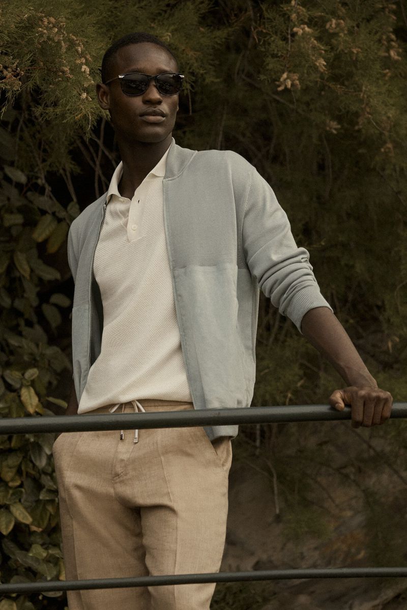 A stylish man stands in muted light, wearing sunglasses, a blue jacket, white polo, and khaki pants, leaning against a railing.
