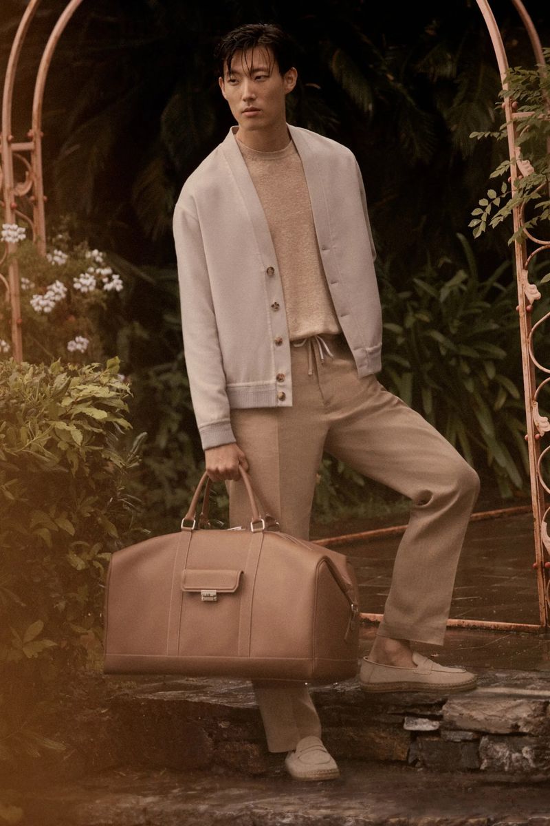 Stylish Asian male model poses outdoors, wearing a beige suit and holding a large leather bag, set against a lush garden backdrop.