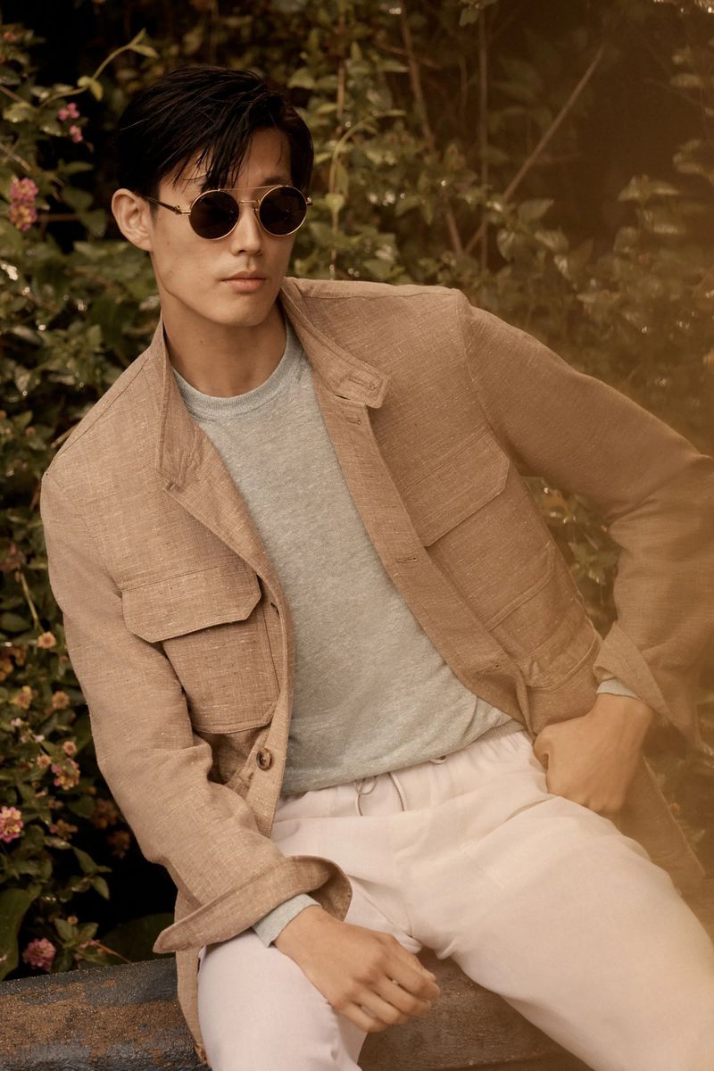 Asian man wearing round sunglasses and a tan jacket sitting outdoors with green foliage background, portrait shot, fashion photography.