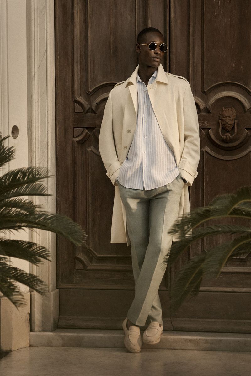 A stylish African-American man in sunglasses and a trench coat poses in front of a brown door with tropical plants to the side.