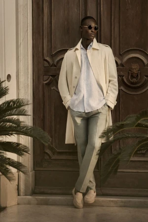 A stylish African-American man in sunglasses and a trench coat poses in front of a brown door with tropical plants to the side.