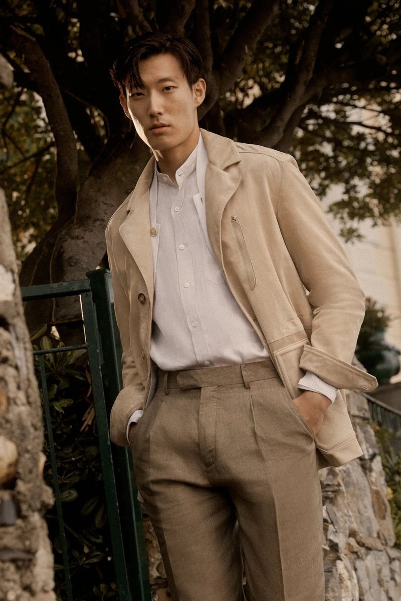 Asian man in beige jacket and trousers poses outdoors against a tree, looking at the camera in a fashion editorial shot.