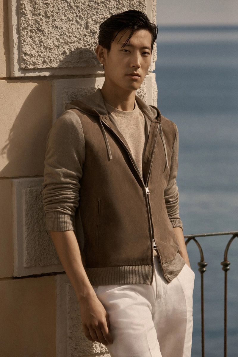 A stylish Asian man in a brown hooded jacket and white pants leans against a wall outdoors, with the ocean in the blurred background.