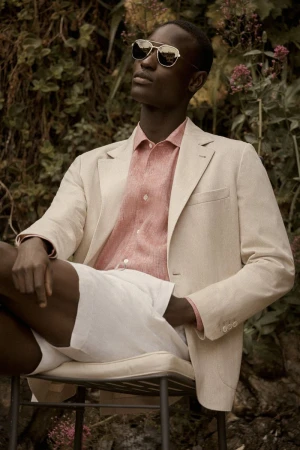 Model wearing sunglasses, beige blazer, and pink shirt sitting outdoors against foliage for Brett Johnson F/W 25