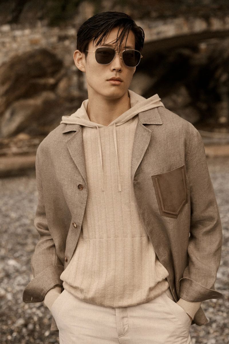Asian man wearing sunglasses, a beige hoodie, and jacket stands outdoors in a fashion pose with hands in his pants pockets.
