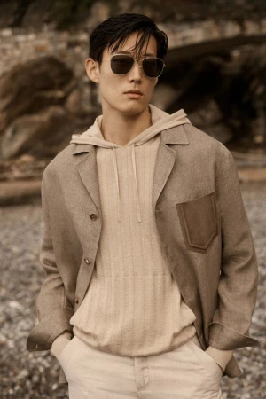 Asian man wearing sunglasses, a beige hoodie, and jacket stands outdoors in a fashion pose with hands in his pants pockets.