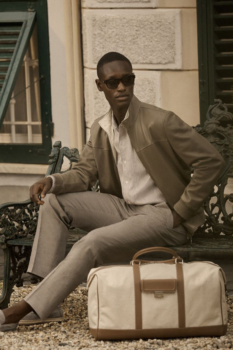 A stylish man in a jacket and sunglasses sits on a bench next to a duffel bag in an outdoor setting with a light neutral color palette.