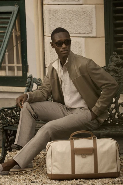 A stylish man in a jacket and sunglasses sits on a bench next to a duffel bag in an outdoor setting with a light neutral color palette.