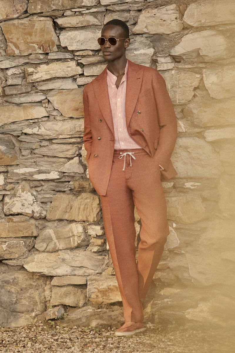 Man in rust-colored suit stands against stone wall, wearing sunglasses and pale pink shirt. Casual yet elegant outdoor fashion.