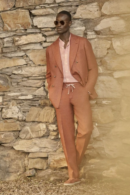 Man in rust-colored suit stands against stone wall, wearing sunglasses and pale pink shirt. Casual yet elegant outdoor fashion.