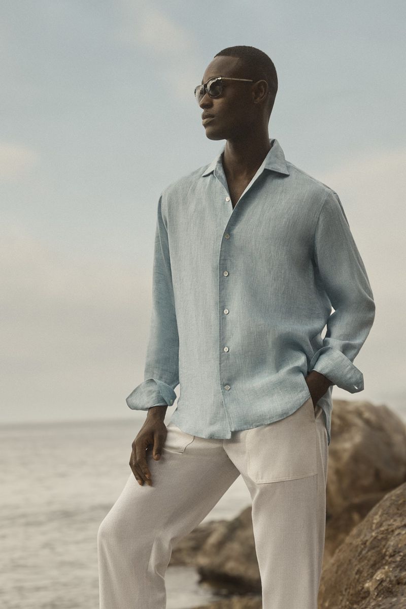 Black model in light blue long-sleeve shirt and white trousers standing on rocky shore by ocean