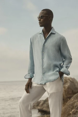 Black model in light blue long-sleeve shirt and white trousers standing on rocky shore by ocean