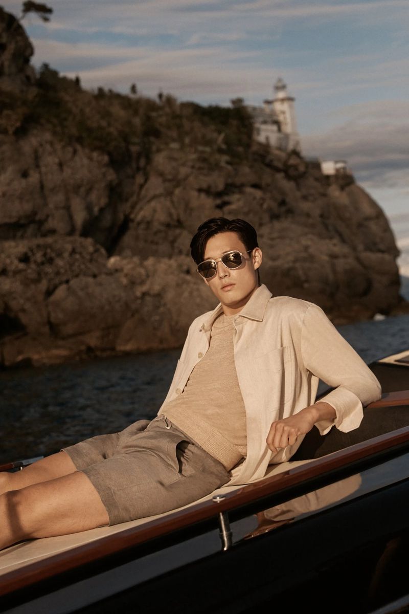 Man poses on boat wearing aviator sunglasses, linen shirt, knit tee, and shorts with a coastal background of rocky cliffs and a building.