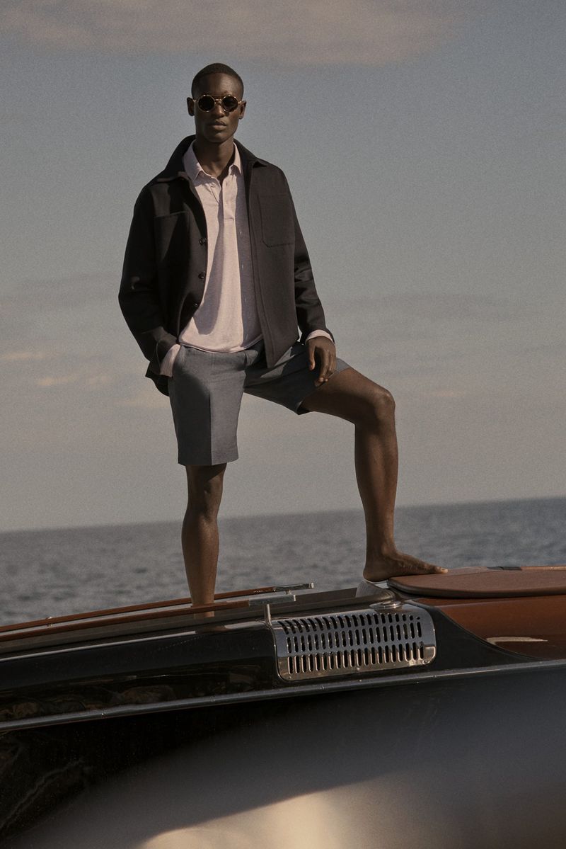Model wearing dark jacket and sunglasses standing on a boat deck over water