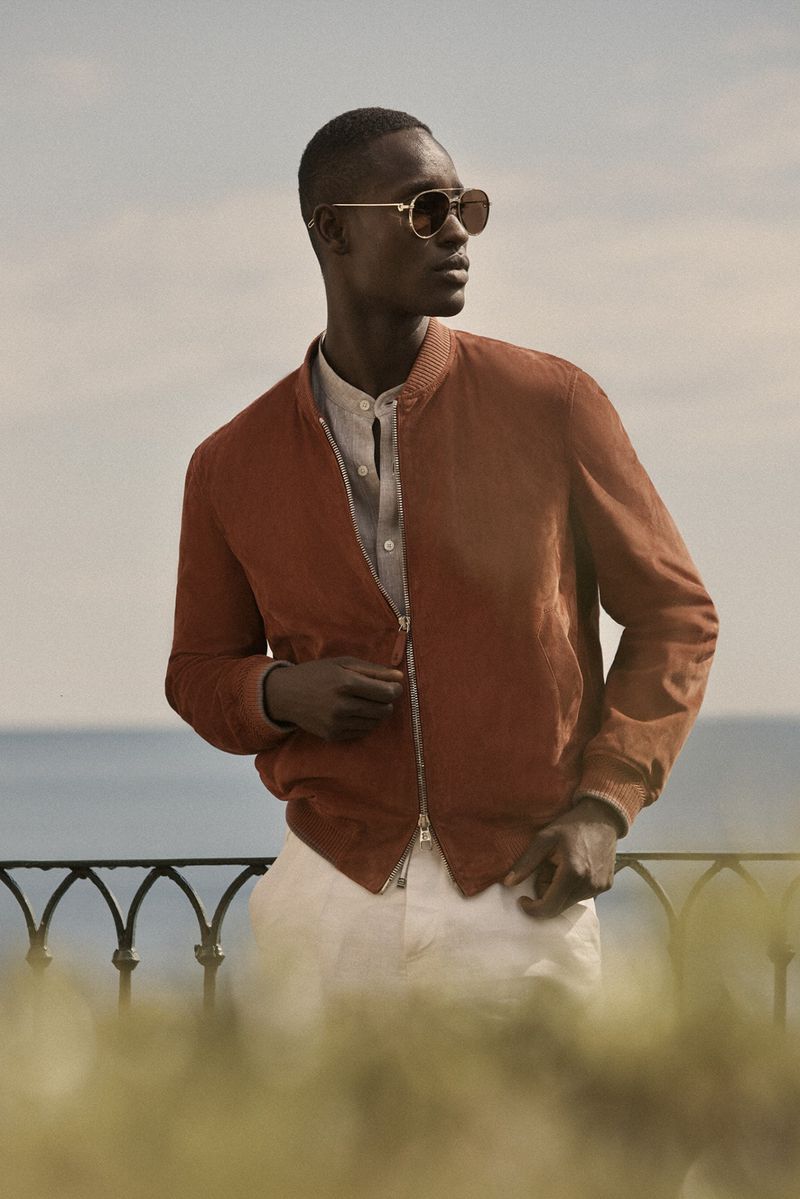 Adult male in outdoor fashion shot. Man in sunglasses and rust-colored jacket stands on a balcony overlooking the sea, blue sky background.