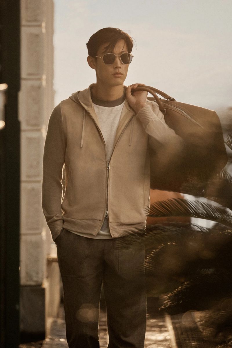 A stylish Asian man wearing sunglasses carries a duffel bag over his shoulder in a relaxed outdoor setting with muted tones.