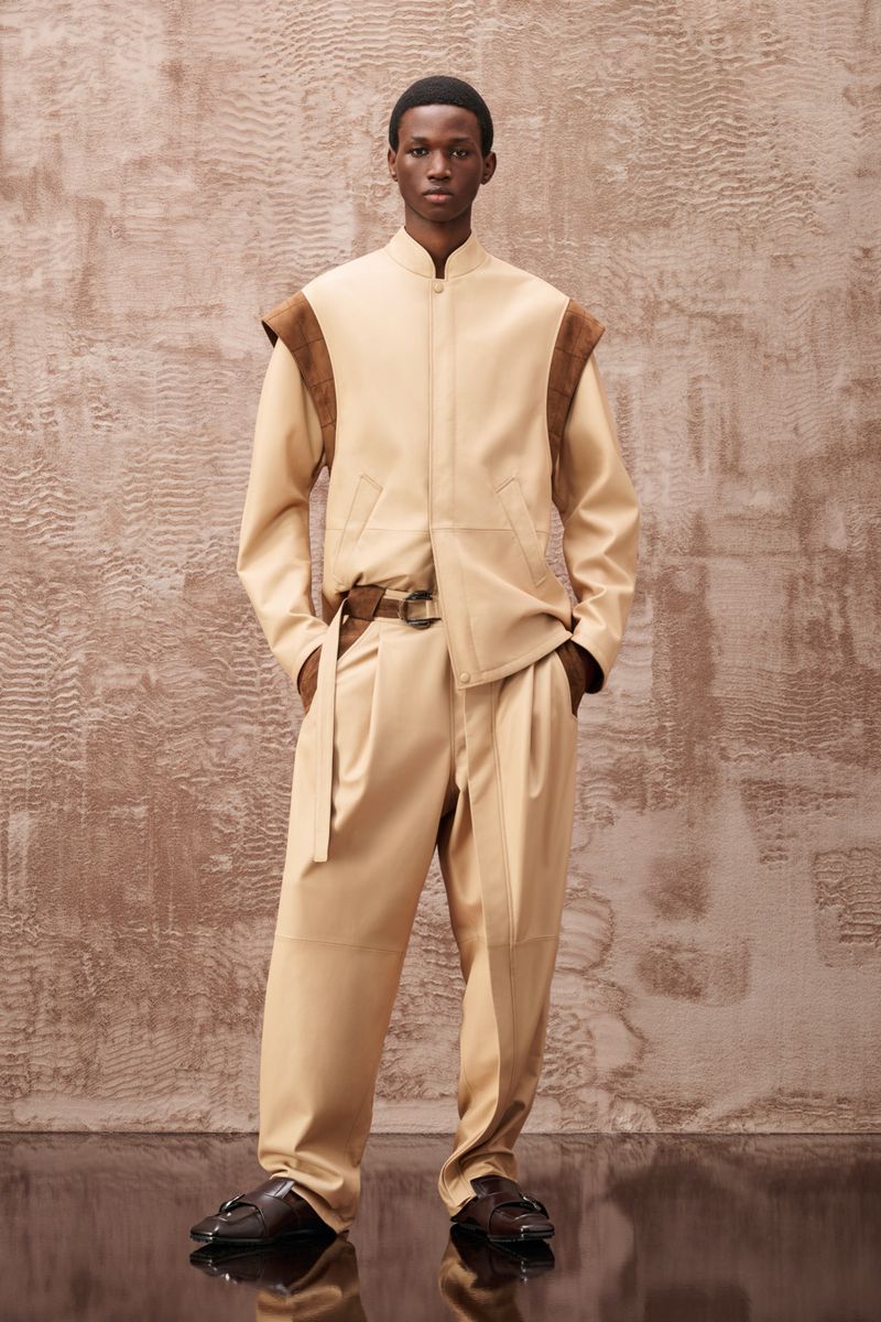 An adult male model poses in a tan-colored outfit featuring a jacket, pants, and belt against a textured background in a studio setting.