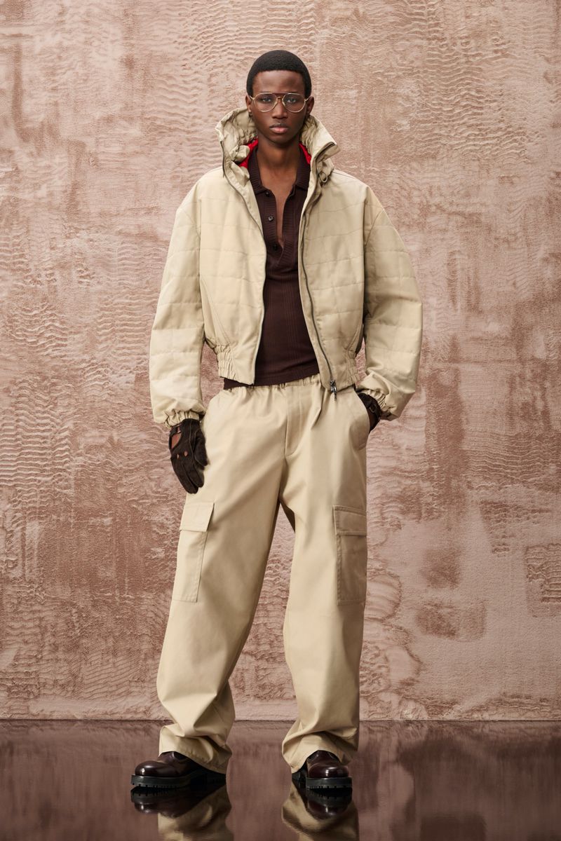 Man in beige jacket and cargo pants, wearing glasses and gloves, stands against textured backdrop in a fashion editorial style.