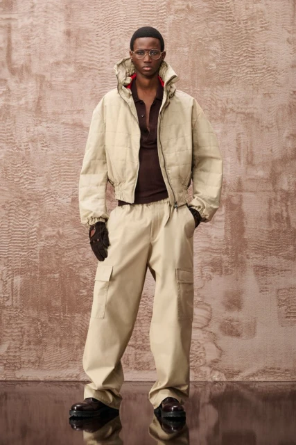 Man in beige jacket and cargo pants, wearing glasses and gloves, stands against textured backdrop in a fashion editorial style.