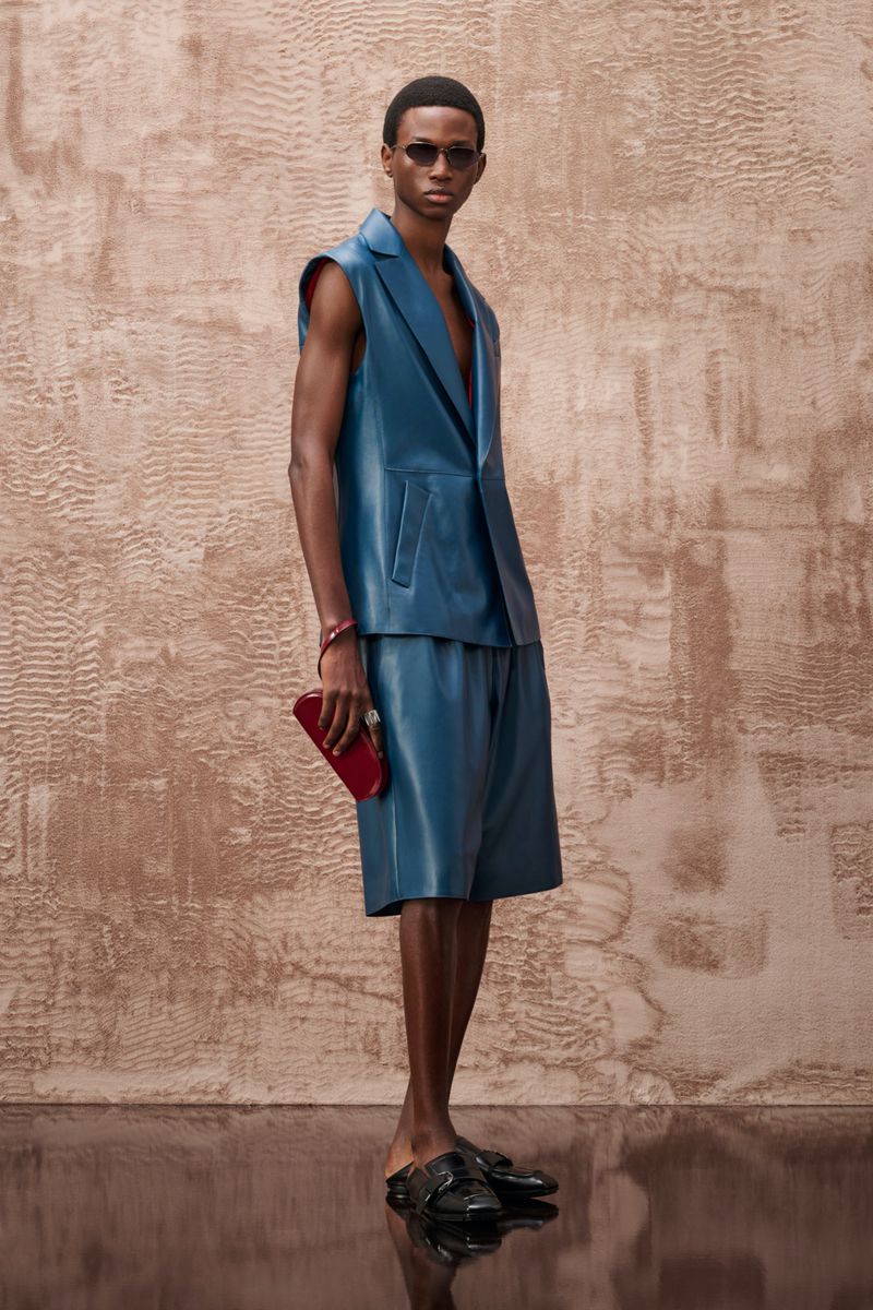 Model wearing a sleeveless blue leather jacket and matching shorts holding a red clutch against a textured wall.