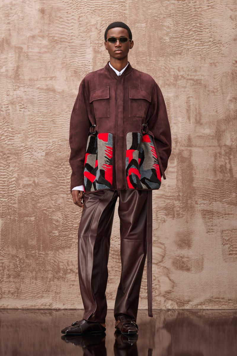 Fashion shot of adult male wearing a brown jacket and leather pants, accessorized with sunglasses and patterned side bags, posing indoors.