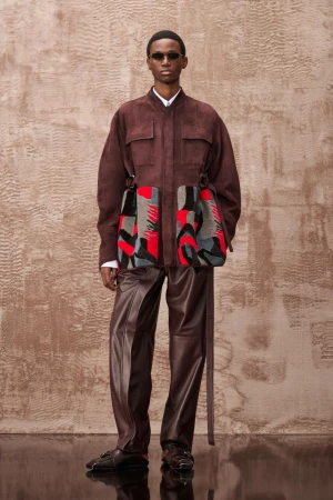 Fashion shot of adult male wearing a brown jacket and leather pants, accessorized with sunglasses and patterned side bags, posing indoors.