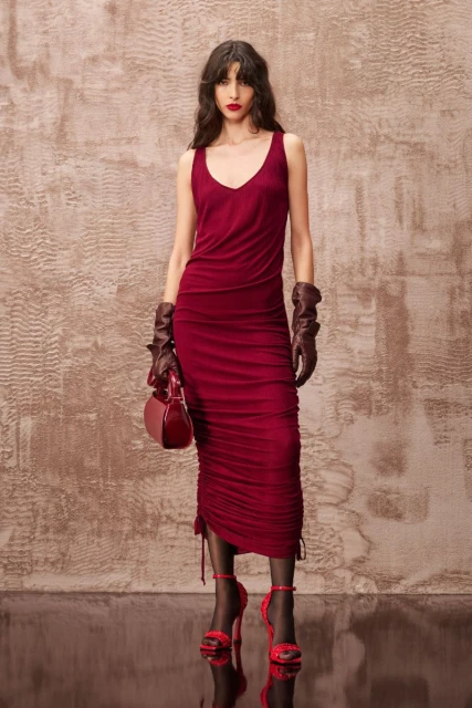Elegant adult female in a burgundy dress, gloves, and heels, carrying a matching purse in a studio setting with textured backdrop.