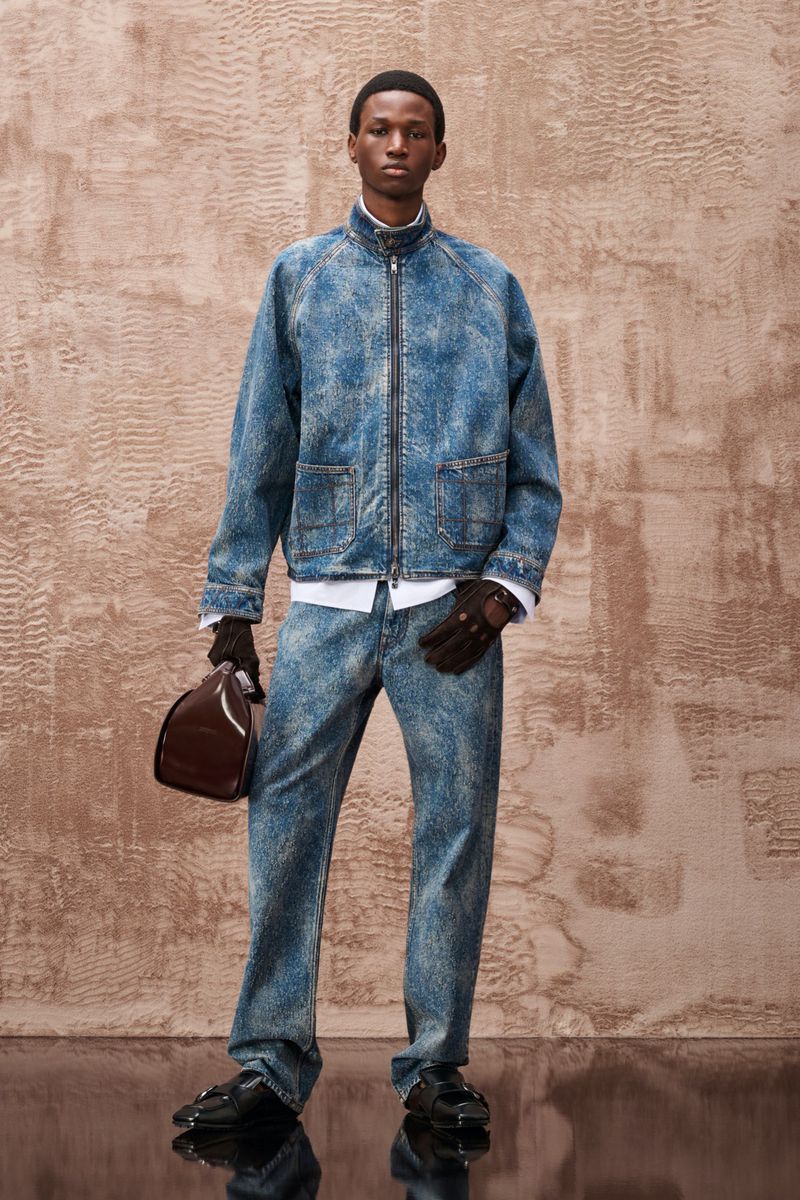 Full-length portrait of a young adult male fashion model wearing a matching acid-wash denim jacket and jeans, holding a helmet.