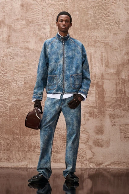 Full-length portrait of a young adult male fashion model wearing a matching acid-wash denim jacket and jeans, holding a helmet.